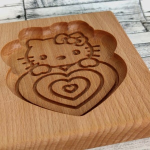 Puede incluir: Cortador de galletas de madera con forma de Hello Kitty sosteniendo un coraz&oacute;n. El coraz&oacute;n tiene c&iacute;rculos conc&eacute;ntricos tallados en &eacute;l.