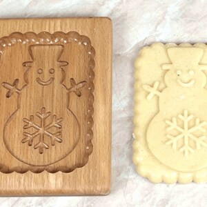 May include: A wooden cookie cutter in the shape of a snowman with a snowflake on its belly. A cookie made with the cutter is shown next to it.