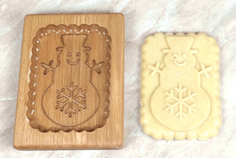 May include: A wooden cookie cutter in the shape of a snowman with a snowflake on its belly. A cookie made with the cutter is shown next to it.