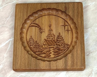 Op de afbeelding: Een houten koekjesvormer met een gedetailleerd ontwerp van een winterscène met een kerk, bomen en een ster. De vormer is vierkant met afgeronde hoeken en heeft een reliëf.