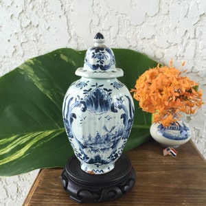 May include: A blue and white ceramic jar with a lid, featuring a landscape design, sits on a black wooden stand. The jar is decorated with floral and architectural patterns. A small vase with orange flowers is in the background.