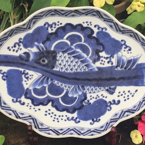 K&ouml;nnte beinhalten: Ein wei&szlig;-blauer Keramikteller mit Fischmuster. Der Teller hat einen gewellten Rand mit einem geometrischen Rand. Der Fisch ist in Blau dargestellt, mit detaillierten Schuppen und Flossen. Der Teller dient wahrscheinlich dekorativen oder Servierzwecken.