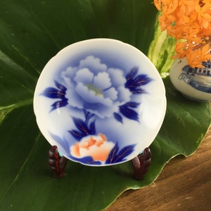 May include: A white and blue porcelain dish with a floral design, on a small wooden stand. The dish features a blue peony with gold accents and an orange flower. The background includes a large green leaf and orange flowers in a vase.