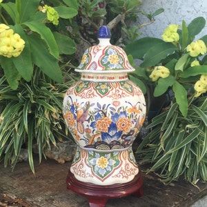 May include: A decorative ceramic ginger jar with a lid, featuring a colorful floral and bird design. The jar is set on a dark wooden stand. The color palette includes blue, orange, green, and white.