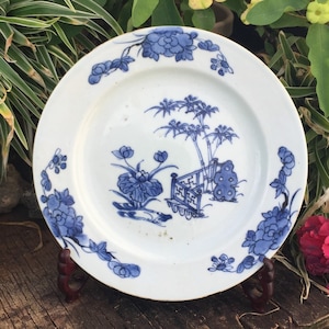 Op de afbeelding: Een wit porseleinen bord met een blauw en wit bloemenmotief. Het bord toont een sc&egrave;ne met een bamboebos, een hek en bloemen. Het bord staat op een houten standaard.