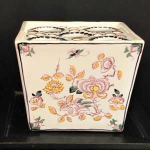 Op de afbeelding: Vierkante keramische bloemenvaas met een crèmekleurige achtergrond en een bloemenontwerp. De bloemen zijn roze en geel met groene bladeren. De bovenkant heeft meerdere gaten voor het rangschikken van bloemen. De vaas heeft een blauwe rand.