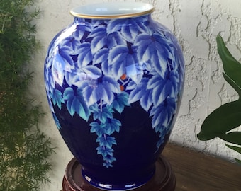 May include: A blue ceramic vase with a gold rim and a dark blue base. The vase features a pattern of blue and white leaves. It sits on a round, dark wooden stand. The vase is a decorative item.