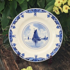 May include: A white ceramic plate with a blue and white design featuring a sailboat on a body of water. The plate has a scalloped edge and a blue and white floral border.