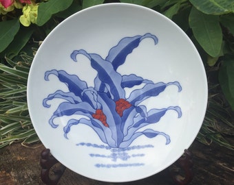 May include: White ceramic plate with a blue and red floral design. The plate features a central cluster of stylized blue leaves and red accents, set against a white background. The plate is displayed on a dark wooden stand.
