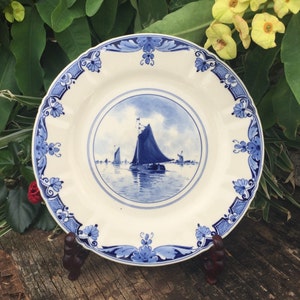 May include: A white ceramic plate featuring a blue sailboat scene. The plate has a decorative blue floral border and a scalloped edge. It is displayed on a dark brown stand, set against a backdrop of green foliage and yellow flowers.