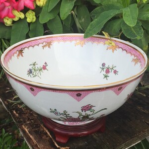 May include: A white porcelain bowl with a pink and gold rim, featuring floral designs. The bowl sits on a dark wooden stand. The rim has a scalloped edge with gold accents. Floral patterns are scattered across the bowl's surface.