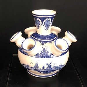 Op de afbeelding: Een wit en blauw Delfts-blauwe tulpenvaas. De keramische vaas heeft een bolvormig lichaam met meerdere openingen voor tulpen en een centrale opening. Het ontwerp omvat blauwe bloempatronen, windmolens en een zeilbootscène.