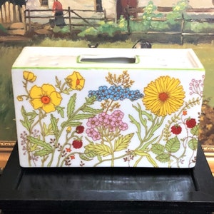 May include: A rectangular white ceramic container with a floral design featuring yellow, blue, and pink flowers, green leaves, and red berries. The container has a rectangular opening on top and a green trim.