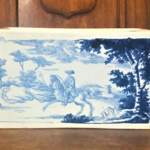 May include: Rectangular white ceramic planter with a blue and white pastoral scene. The design shows a person on horseback, trees, and a building. The planter has a slightly distressed appearance.