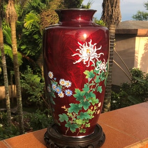 May include: A tall, red decorative vase with a floral design. The vase features white chrysanthemums and blue and white flowers with green leaves. The vase is on a dark brown wooden stand.