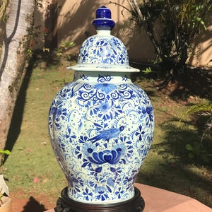May include: A large, ornate blue and white ceramic jar with a lid. The jar features a detailed floral and bird pattern. It is displayed on a dark wooden stand, set against a backdrop of greenery.