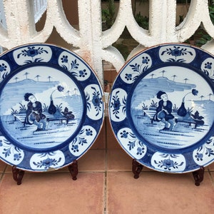 Op de afbeelding: Twee blauwe en witte keramische borden met een decoratieve rand en een scène met een persoon die op een bankje in een landschap zit. De borden staan op houten standaard.