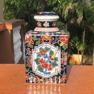 Könnte beinhalten: Eine quadratische Keramikdose mit Deckel, verziert mit einem floralen Muster in Rot-, Blau-, Weiß- und Gelbtönen auf schwarzem Hintergrund. Die Dose hat ein traditionelles asiatisches Design.