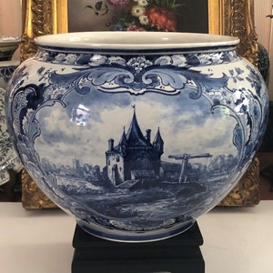 Op de afbeelding: Een grote, blauw-witte Delftse pot met een afbeelding van een Nederlands dorp en een windmolen. De pot is versierd met ingewikkelde bloemmotieven en staat op een zwarte houten standaard.