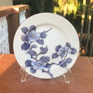 May include: White ceramic plate with a floral design in shades of blue and gold. The plate features a branch with blue flowers and leaves. The plate is displayed on a clear plastic stand, showcasing the decorative tableware.