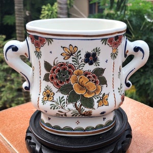 Op de afbeelding: Een witte keramische plantenbak met twee handvatten, versierd met een bloemmotief in geel, rood en groen. De plantenbak staat op een donkere houten standaard, die een traditionele, decoratieve stijl laat zien. Het bloemmotief bevat gedetailleerde bladeren en bloemen.