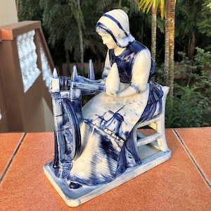K&ouml;nnte beinhalten: Blau-wei&szlig;e Keramikfigur, die eine Frau an einem Spinnrad darstellt. Die Figur hat ein traditionelles niederl&auml;ndisches Design, mit einer Frau, die eine Haube und eine Sch&uuml;rze tr&auml;gt. Die Frau sitzt auf einem Stuhl.