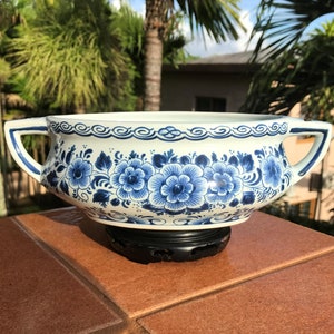 Op de afbeelding: Een witte keramische kom met blauwe bloemmotieven en twee handvatten. De kom is versierd met blauwe bloemen en bladeren. Hij staat op een zwarte standaard. De achtergrond omvat groene bomen en een blauwe lucht.