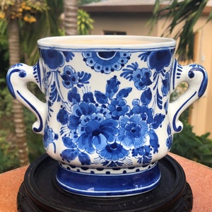 Op de afbeelding: Een wit en blauw keramieken plantenbak met twee handvatten, versierd met een bloemmotief. De plantenbak is versierd met blauwe bloemen en bladeren op een witte achtergrond. Hij staat op een donkere houten standaard.