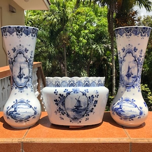 May include: Three-piece ceramic set with a Delftware design. The set includes two tall vases and a rectangular planter. Each piece features a white background with blue floral patterns and scenic illustrations of sailboats and windmills.