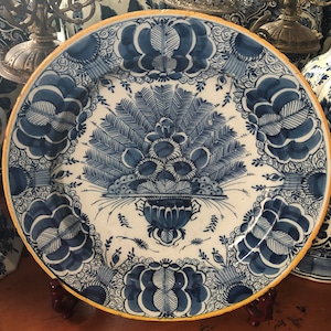 Puede incluir: Un plato redondo de cerámica blanca con un diseño floral azul. El plato presenta un motivo central de un arreglo floral estilizado y un borde de patrones florales. El borde está delineado en amarillo. Vajilla decorativa.