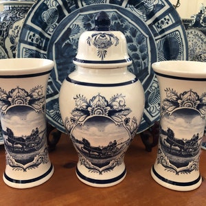 May include: A collection of white and blue ceramic vases and decorative plates. The vases feature a landscape scene with a horse-drawn carriage. The plates and vases have a traditional Delftware design.