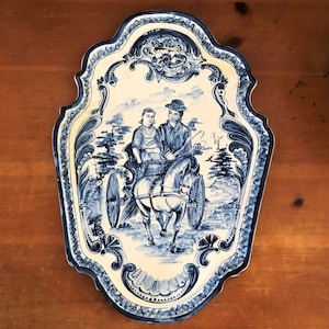 Op de afbeelding: Een decoratieve keramische serveerschaal met een blauw en wit ontwerp. De schaal toont een gedetailleerde illustratie van een paardenkoets met twee figuren. De schaal heeft een geschulpte rand en een sierlijke rand, wat een vleugje elegantie toevoegt.
