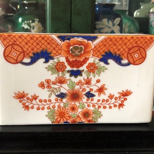 Op de afbeelding: Rechthoekige witte keramische container met oranje, blauwe en groene bloemen- en geometrische patronen. De container is versierd met een centraal bloemmotief en symmetrische patronen aan de bovenkant. Het staat op een zwarte houten standaard.