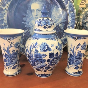 May include: A collection of white and blue ceramic vases and a lidded jar. The pieces feature a floral design in shades of blue. The vases have a flared shape, and the jar has a bulbous body. Decorative plates are in the background.