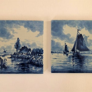 Puede incluir: Dos baldosas de cer&aacute;mica cuadradas con pinturas de paisajes azules y blancas. Una baldosa representa una escena rural con una casa, &aacute;rboles y un molino de viento. La otra muestra veleros en el agua bajo un cielo nublado.