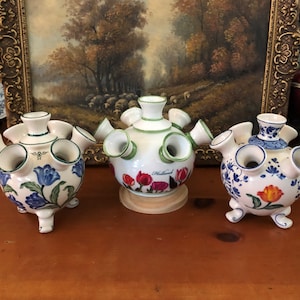 May include: Three ceramic tulip vases with multiple openings for flowers. The vases are white with hand-painted floral designs in blue, red, and orange. Each vase has a unique shape and design, with a wooden base on one.