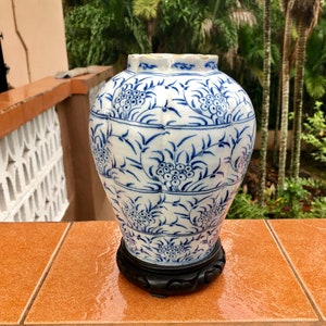 Op de afbeelding: Een blauwe en witte porseleinen vaas met een bloemenmotief. De vaas staat op een houten standaard.