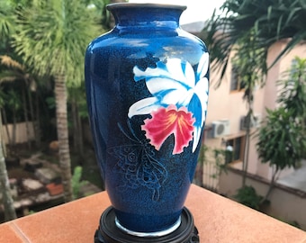 May include: A blue ceramic vase with a floral design featuring white, red, and blue flowers. The vase is set on a dark wooden stand. The background shows a blurred outdoor scene with trees and buildings.