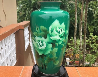 May include: A green ceramic vase with white floral designs. The vase is on a black wooden stand with three legs.
