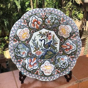 K&ouml;nnte beinhalten: Ein gro&szlig;er, runder, wei&szlig;er Teller aus Keramik mit einem gewellten Rand. Der Teller ist mit einem gr&uuml;nen, blauen und orangefarbenen Blumenmuster und einem Pfauenmotiv in der Mitte verziert.