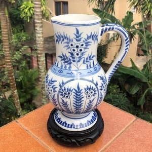 Op de afbeelding: Een witte keramische kan met een blauw en wit bloemenmotief. De kan heeft een rond lichaam en een handvat. Hij staat op een houten voet.