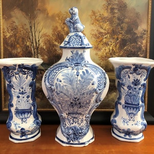 May include: A set of three Delftware ceramic pieces, featuring a large lidded vase with a squirrel finial and two matching candle holders. The blue and white hand-painted floral designs are characteristic of Delft pottery.