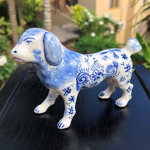 Puede incluir: Figura de perro de cer&aacute;mica en blanco y azul. El perro tiene orejas azules y una cabeza salpicada de azul. El cuerpo es blanco con patrones florales y geom&eacute;tricos azules. La figura est&aacute; de pie sobre cuatro patas con una peque&ntilde;a cola.