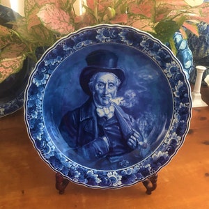 K&ouml;nnte beinhalten: Ein blauer und wei&szlig;er Keramikteller mit einem erh&ouml;hten Rand und einem Blumendesign. In der Mitte des Tellers befindet sich ein Portr&auml;t eines Mannes in einem blauen Mantel und einem schwarzen Hut, der eine Pfeife h&auml;lt.