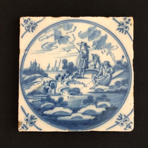 May include: Square ceramic tile featuring a hand-painted blue and white pastoral scene. The scene shows figures fishing and herding animals near water, with trees and buildings in the background. Decorative floral corner accents.