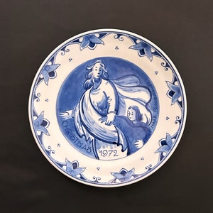 Puede incluir: Un plato de cerámica azul y blanco con una escena navideña. El plato presenta a una mujer con una túnica azul sosteniendo a un niño. El texto "Christmas 1972" está escrito en el plato.