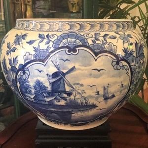 May include: A large, round, white and blue ceramic planter. The planter features a detailed blue and white windmill scene, surrounded by floral patterns. The rim has a decorative blue border. It is placed on a black stand.