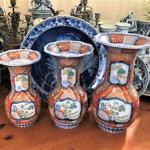 May include: Three antique Japanese vases with a red and orange floral design. Each vase features a landscape scene in a white framed panel. The vases are displayed with other blue and white porcelain pieces, typical of the period.