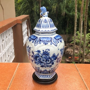 May include: A white and blue ceramic vase with a lid, featuring floral patterns. The vase has a black base and a decorative finial on top. The design is in a classic blue and white color scheme.