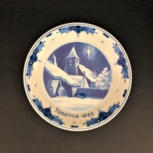 May include: A blue and white ceramic plate featuring a winter scene of a church and houses. The plate has a blue border with a holly leaf design and the text "Kerstmis 1962".